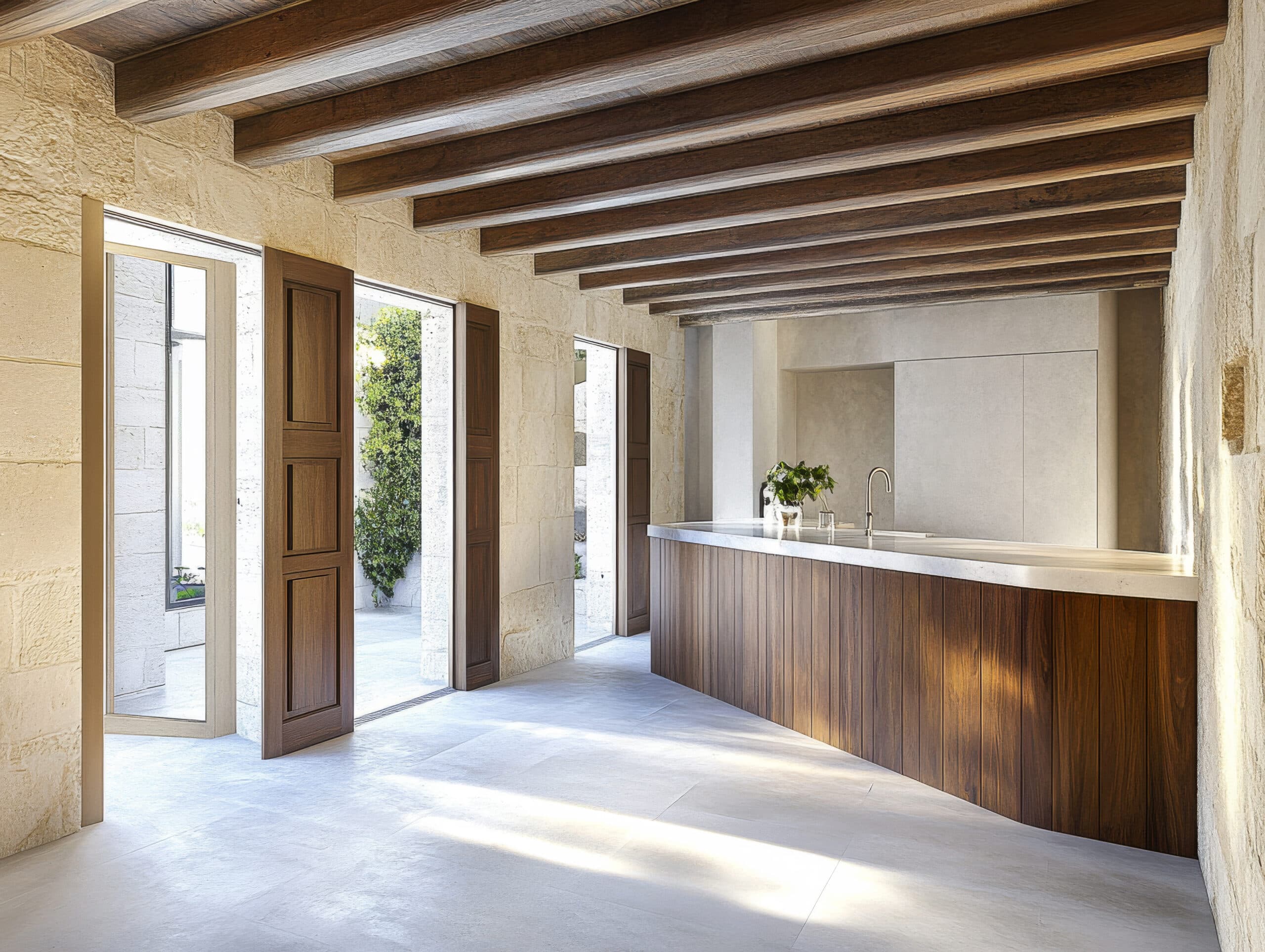 A spacious kitchen features wooden beams overhead and a sleek countertop. Sunlight floods through large windows and doors, creating an inviting atmosphere for cooking and dining.